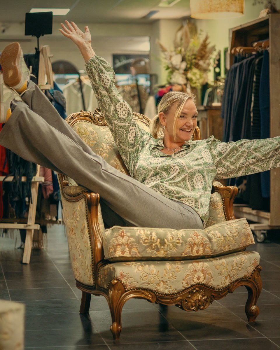 Nance zit lachend in een antieke stoel tussen de kledingrekken van haar winkel